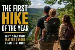 Father carrying toddler in a hiking backpack while his wife walks beside him on a forest trail overlooking a river valley during their first family hike of the year.